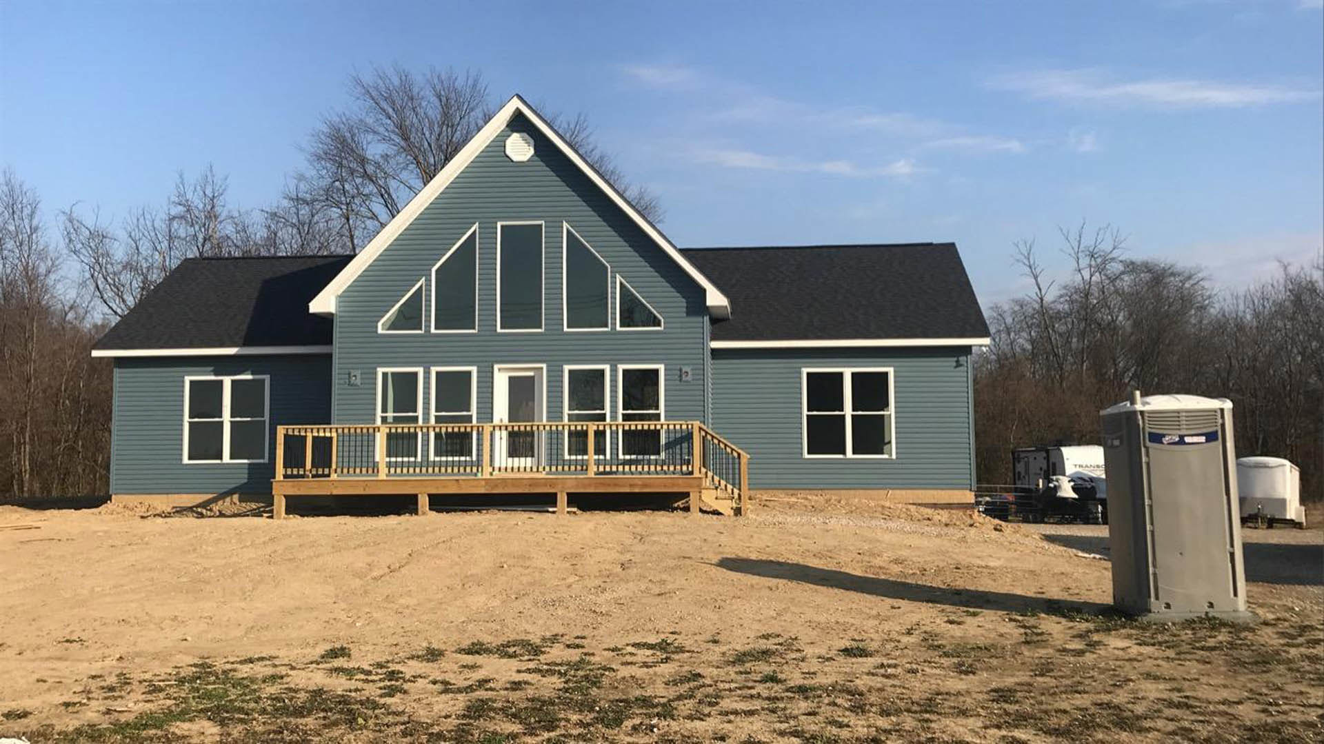 Two-story house with white siding, covered front porch with wooden railing, fenced yard, portable toilet on dirt patch, scattered grass, large windows, cloudy sky.