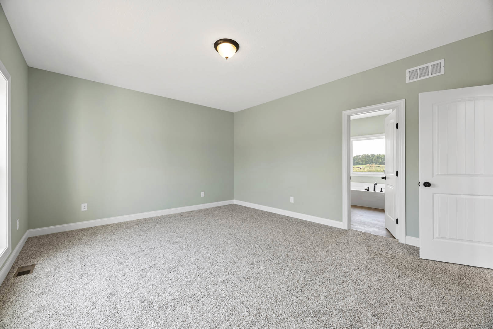 Carpeted room with white walls, white door, ceiling light fixture, wall vent, and adjacent bathroom featuring a window and bathtub