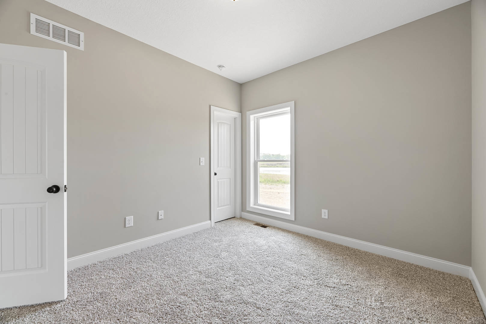 White paneled door with black handle, beige carpeted floor, white baseboards, wall vent, window overlooking river, smooth plaster walls