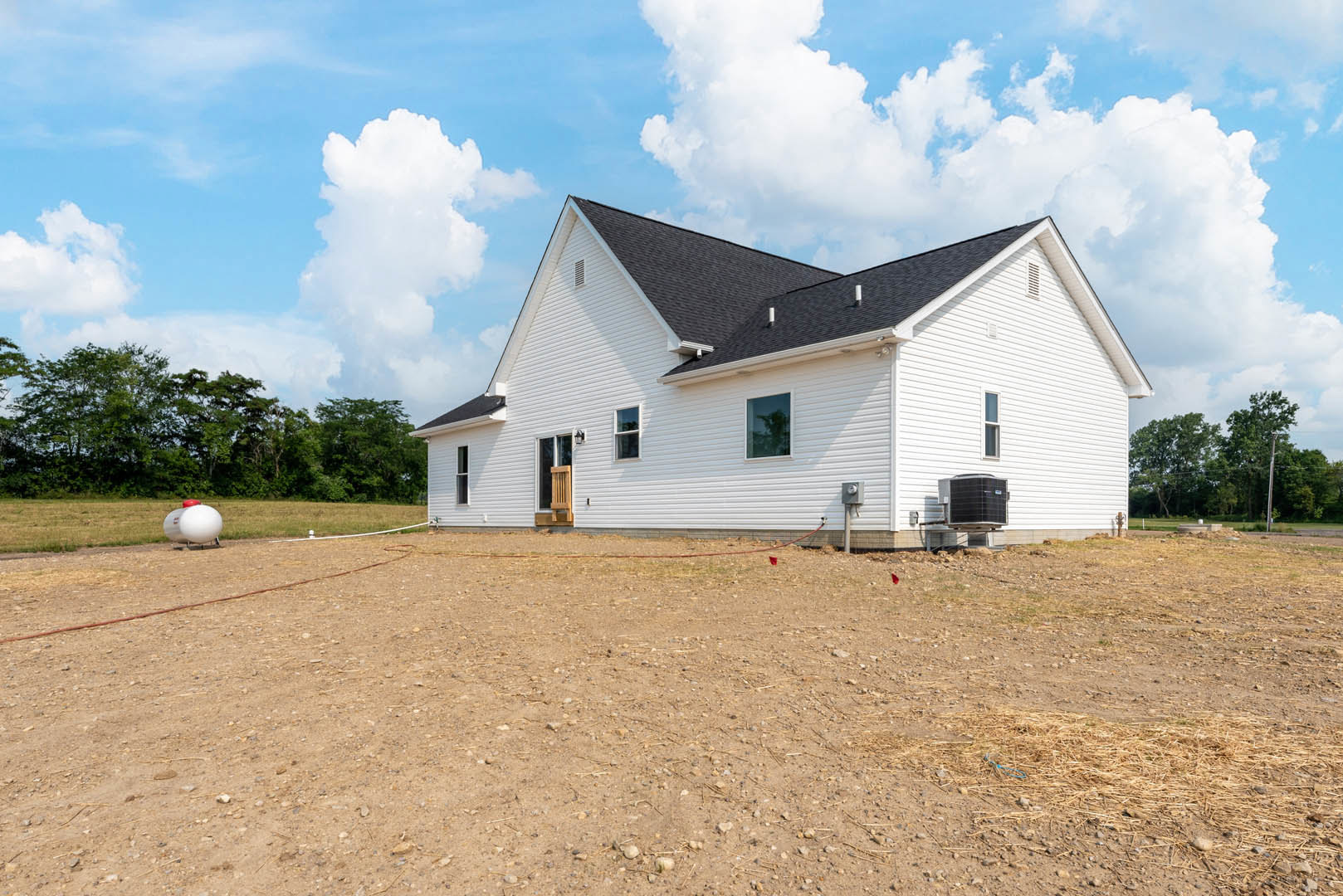 White house with black roof, dirt field in foreground, scattered trees and cloudy sky in background, large white cylinder with red cap near house, window reflecting tree, large