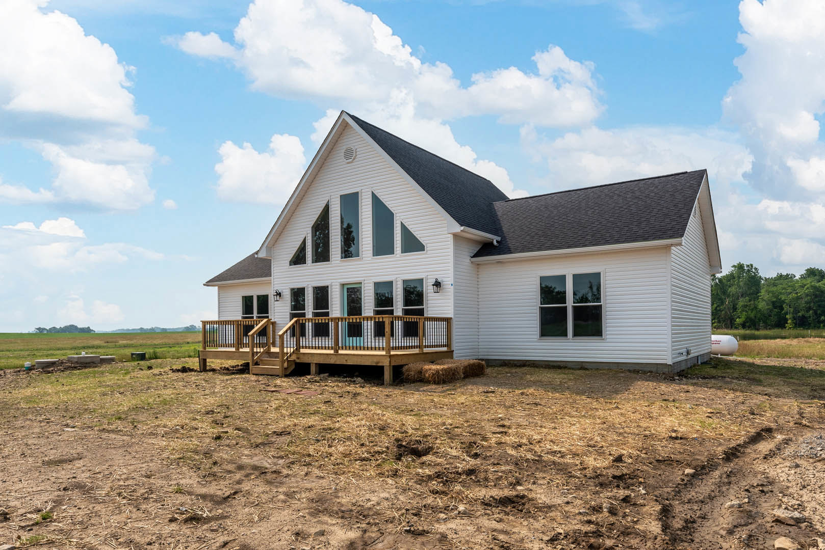 Wooden deck with white railing overlooking a grassy field, large windows with white frames, hay bales stacked near the deck, cottage-style house exterior with light siding and