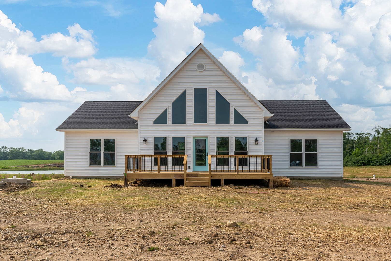 White cottage-style house with wooden deck, blue front door, white railing, and steps, set against a partly cloudy sky with dirt and grass in the yard.