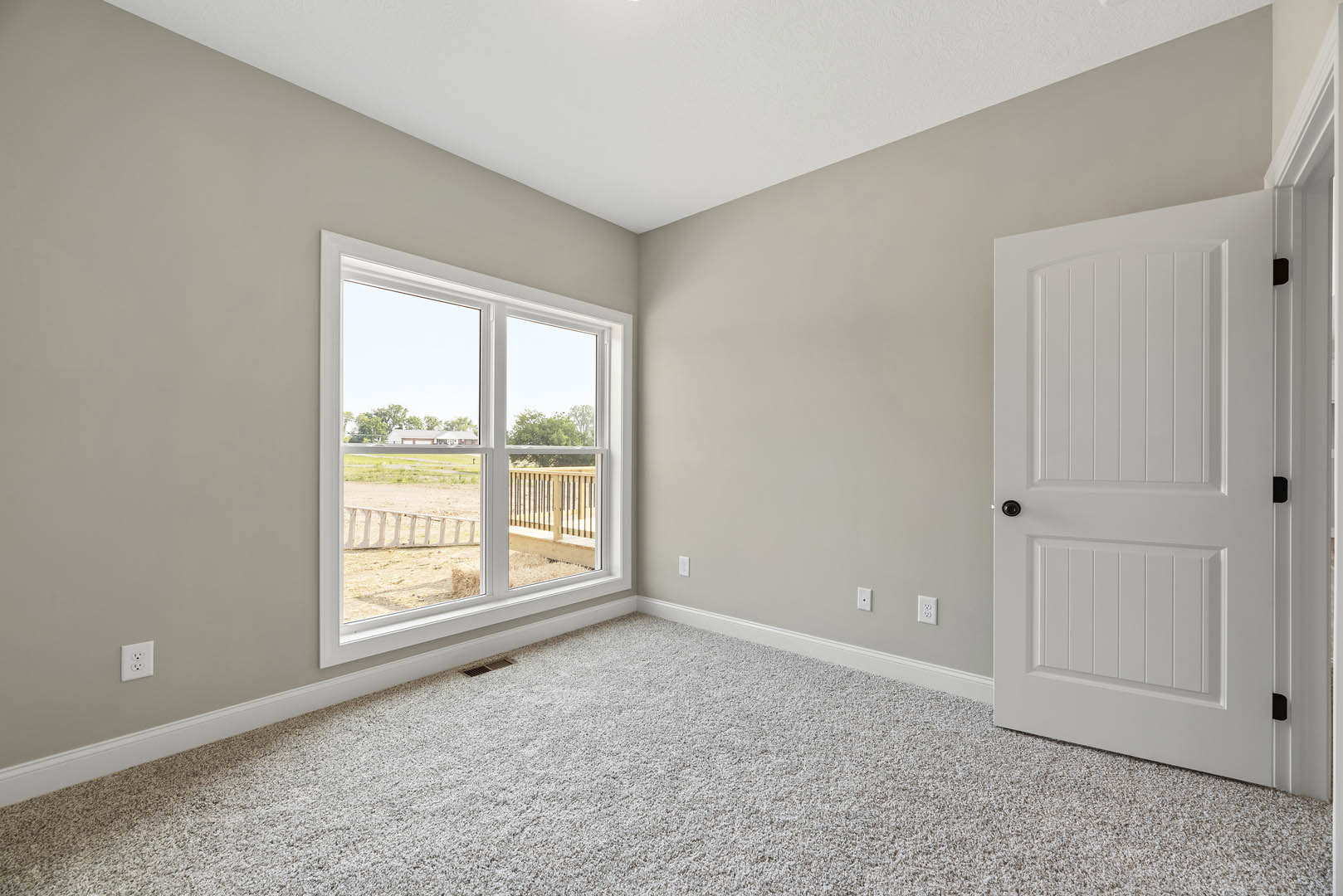 White carpeted room with a white door featuring a black knob, a window overlooking a field and fence, a white electrical outlet on the wall, and a person standing near the white