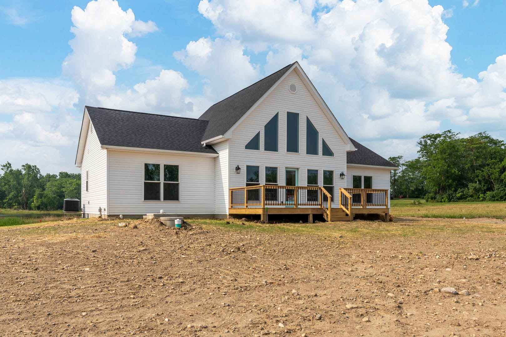 Two-story house with light siding, large wooden deck with railing, surrounded by grassy yard and mature trees under partly cloudy sky
