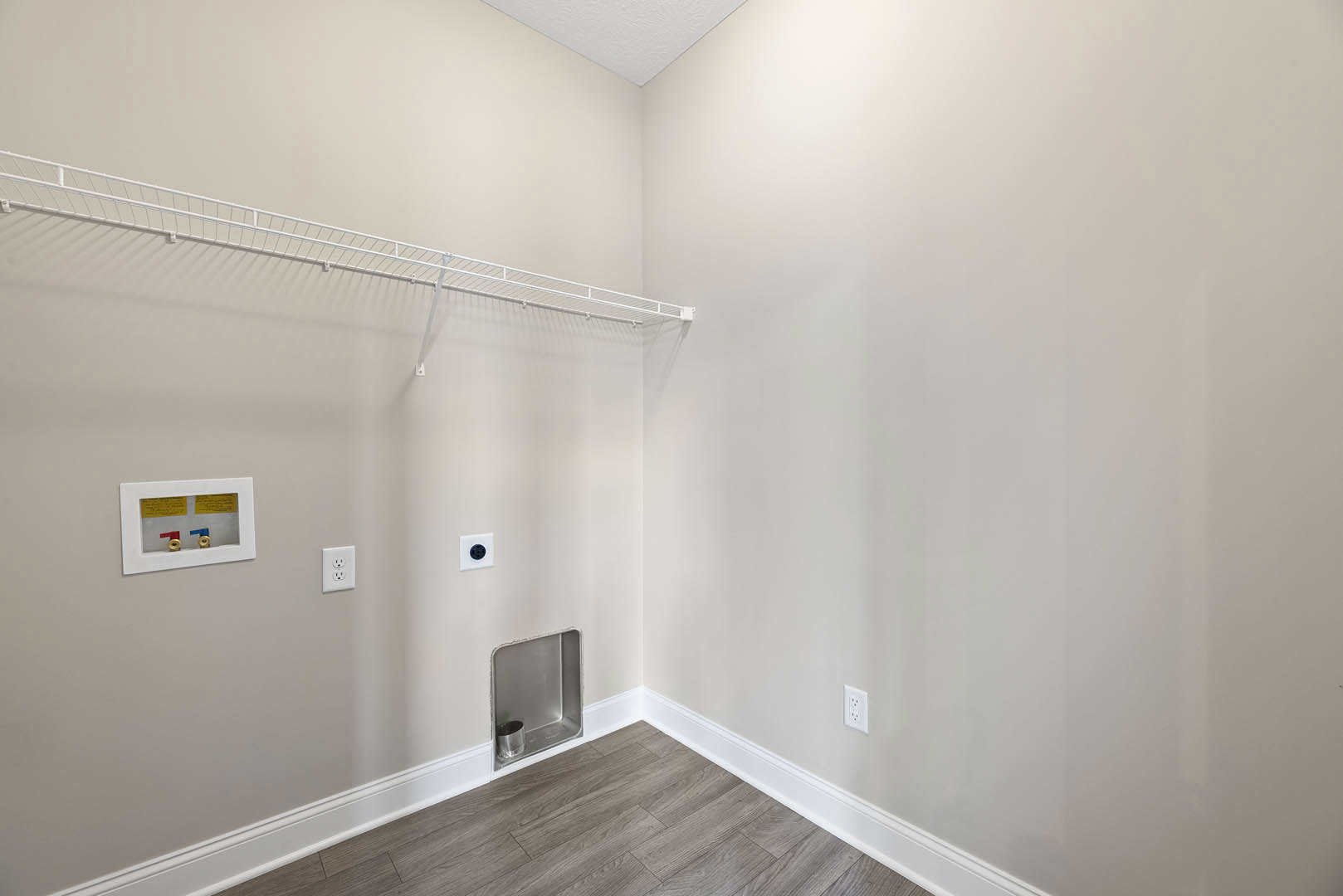 White plaster wall with built-in white shelf, white metal rack mounted on wall, white outlet with black plugs, window framed in white with colored pipes visible, metal container