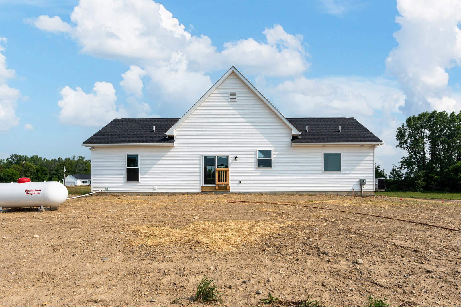 White house with wooden door and vent, surrounded by dirt field, trees in background, white tank with red text nearby