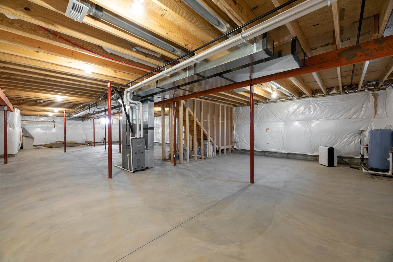 Unfinished basement with exposed wood framing, white plastic vapor barrier on walls, blue and white plumbing pipes, grey rectangular HVAC unit with black vents, and white