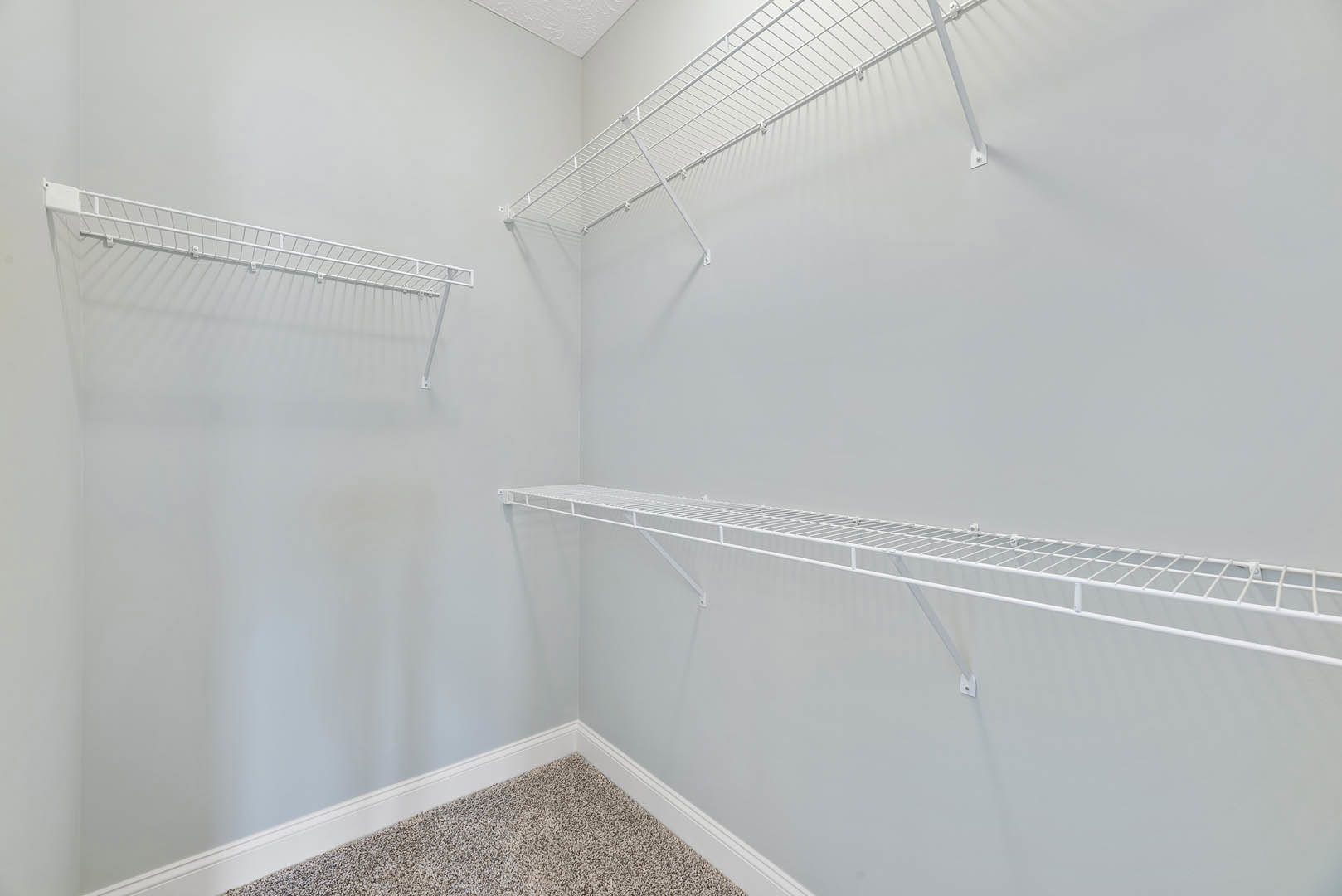 White wire shelving mounted on a white wall above light carpet flooring in a bathroom with plaster ceiling and tiled shower visible.
