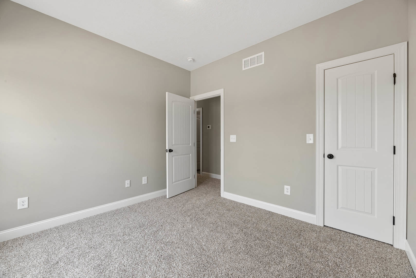 White paneled doors with black knobs, beige carpet flooring, white electrical outlet on wall, ceiling vent, smooth plaster walls