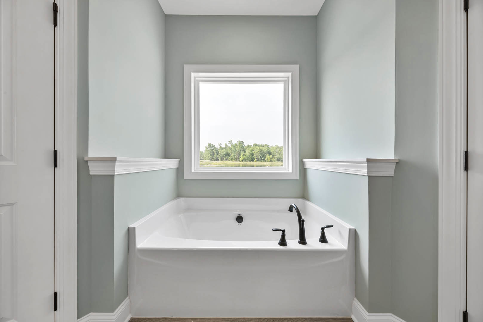 White freestanding bathtub with black faucet beneath large window overlooking green trees, surrounded by light tile walls and minimalist bathroom fixtures