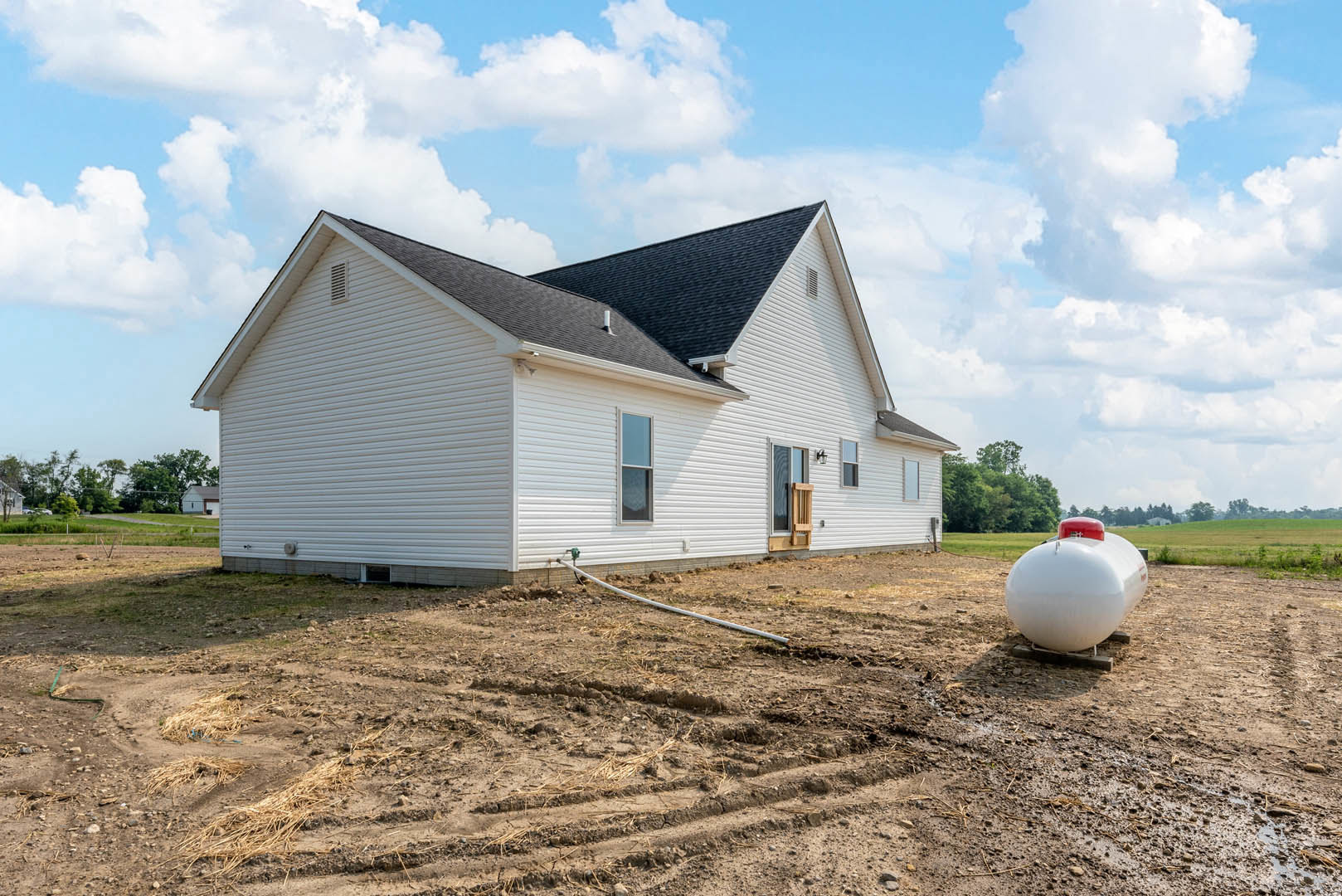 White house with black roof, large white tank with red top in front, pipe lying on dirt ground, grassy yard, cloudy sky overhead
