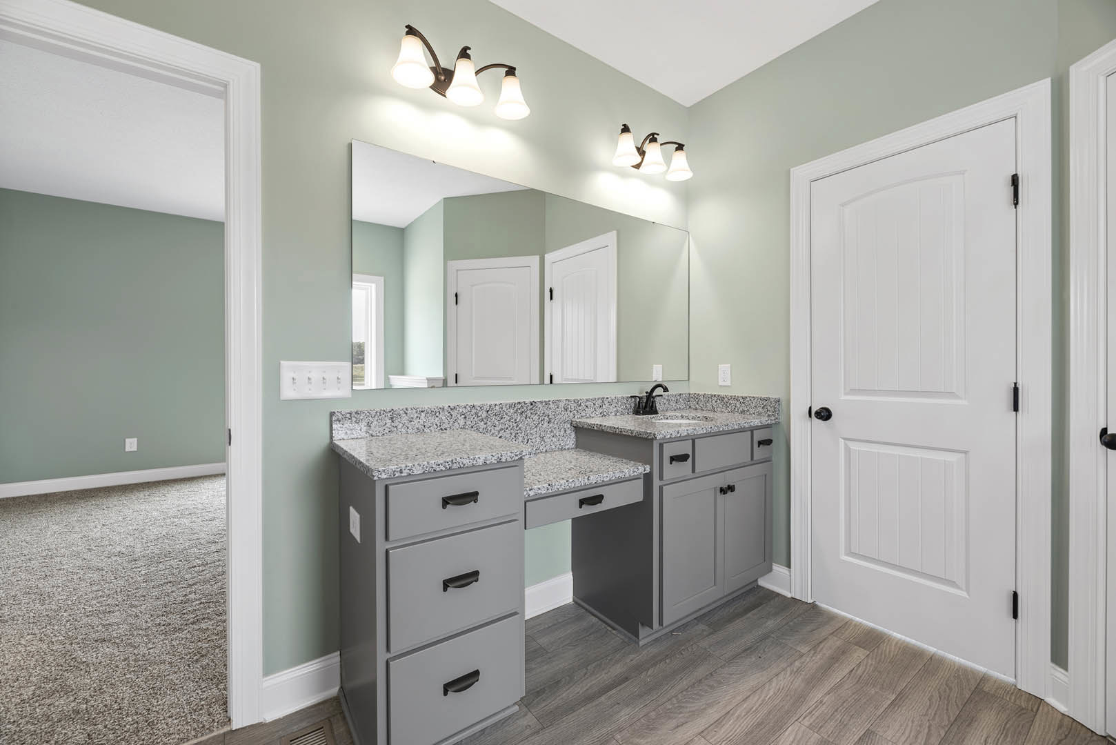 Bathroom with grey vanity cabinet and black handles, white countertop with black faucet, large mirror above, white door with black knobs, tile flooring, close-up of light fixture