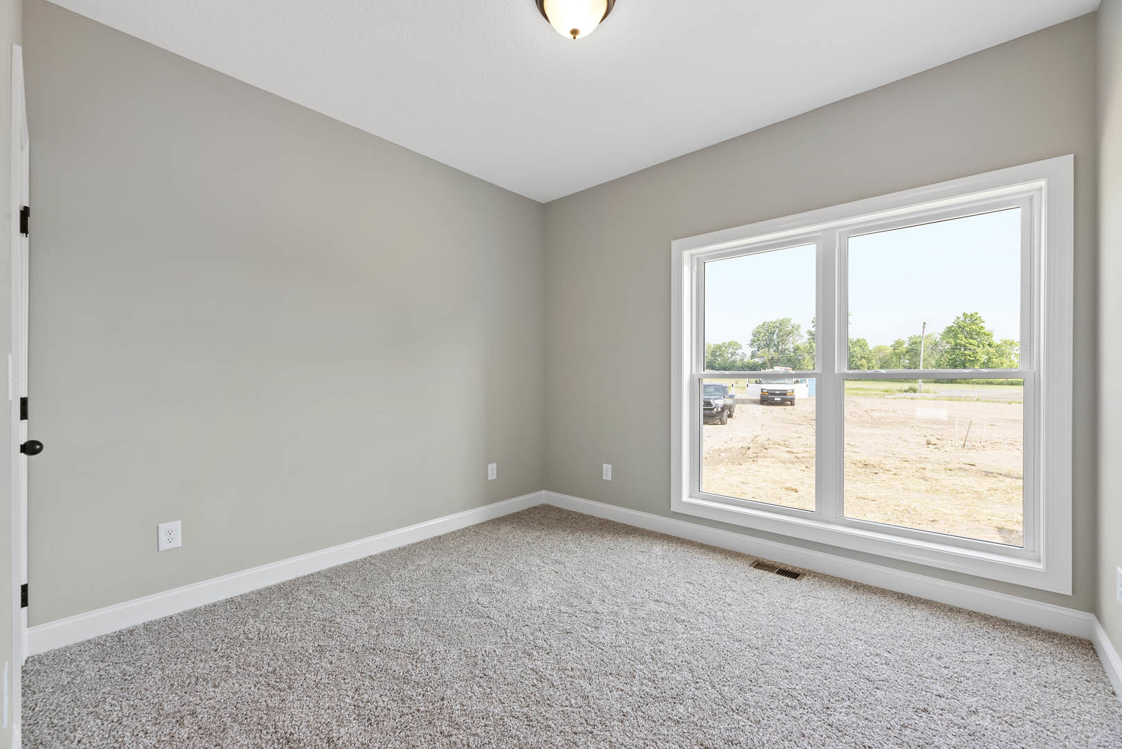 Bedroom with beige carpet, white walls, ceiling light fixture, and large window overlooking dirt field and parked cars
