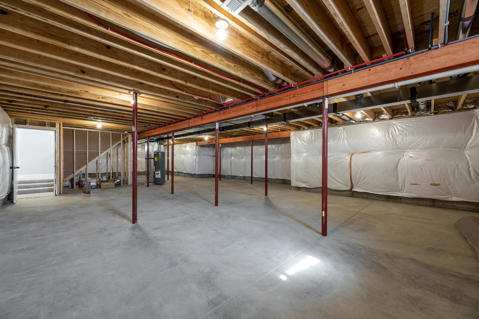 Room with exposed wooden ceiling, prominent steel beam, concrete floor, red support poles, white wall covering, and illuminated light fixture