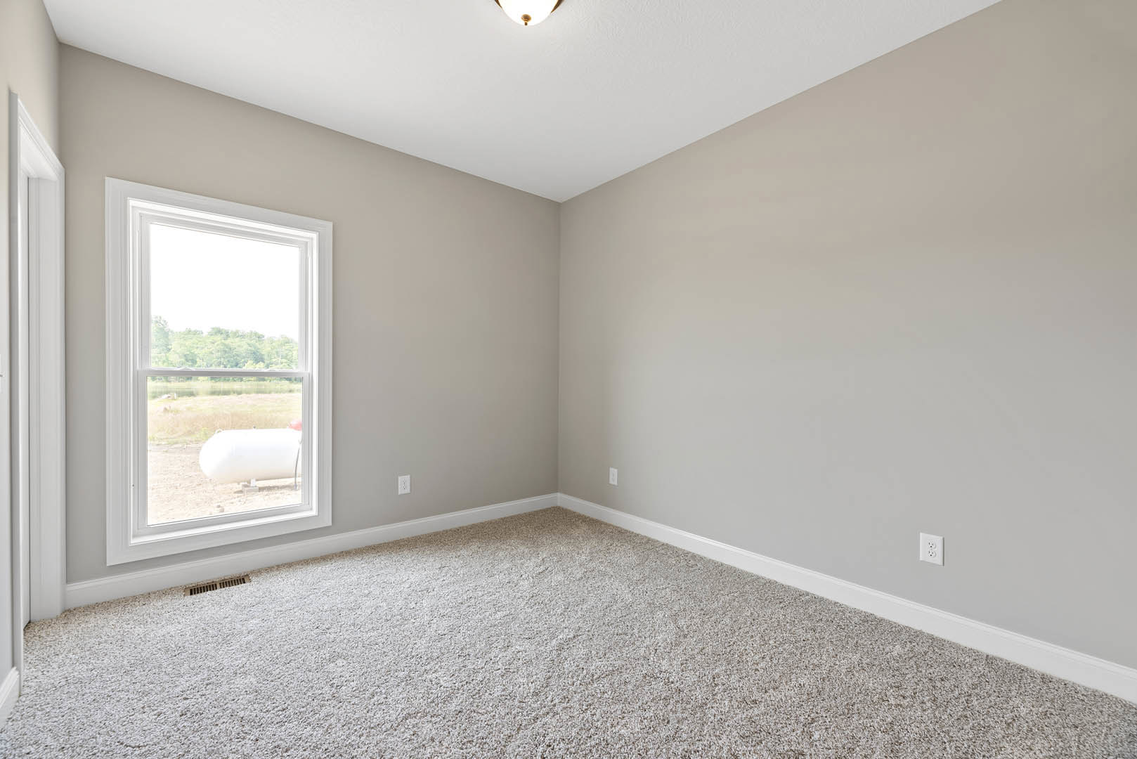 Carpeted room with white walls, large window overlooking a white tank outside, ceiling-mounted light fixture, and simple molding along the ceiling.