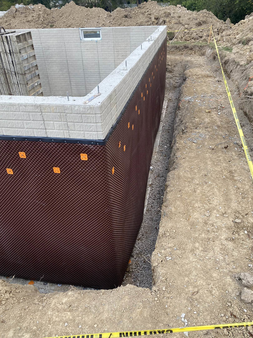 Partially built brick and concrete exterior wall with white-framed window, stack of wooden planks, and yellow tape marking ground at residential construction site