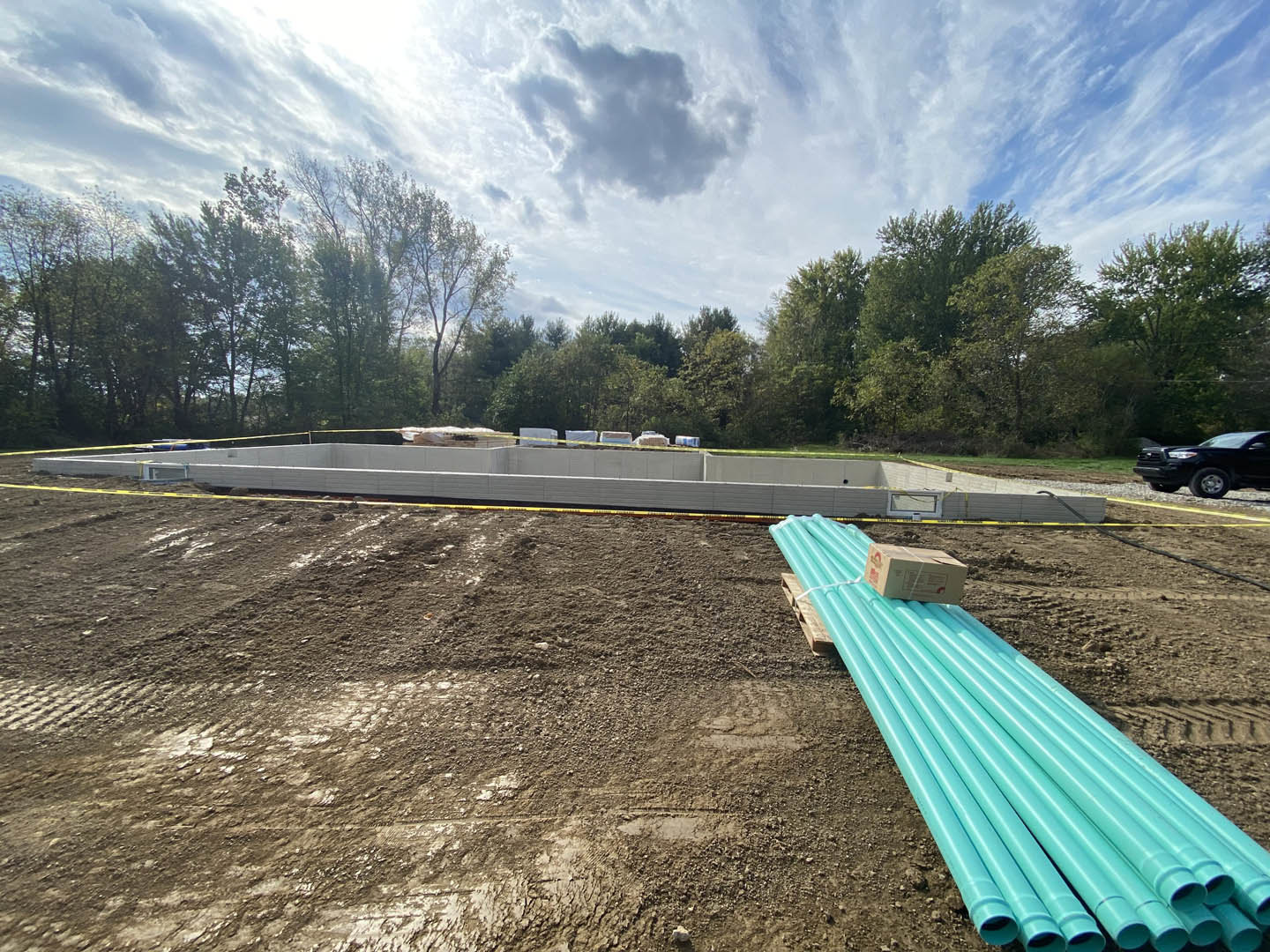 Blue PVC pipes stacked on a wooden pallet beside a pile of cement on a dirt construction site, with yellow caution tape, scattered materials, and trees under a cloudy sky.