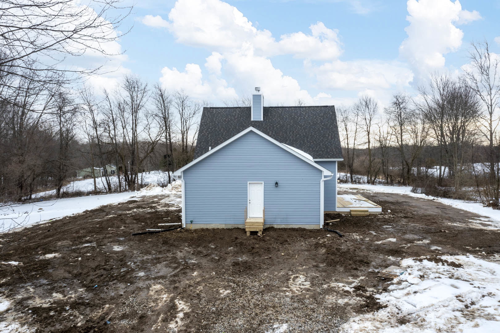 Blue house with white door and blue siding under construction, surrounded by snow and bare trees, cloudy winter sky overhead
