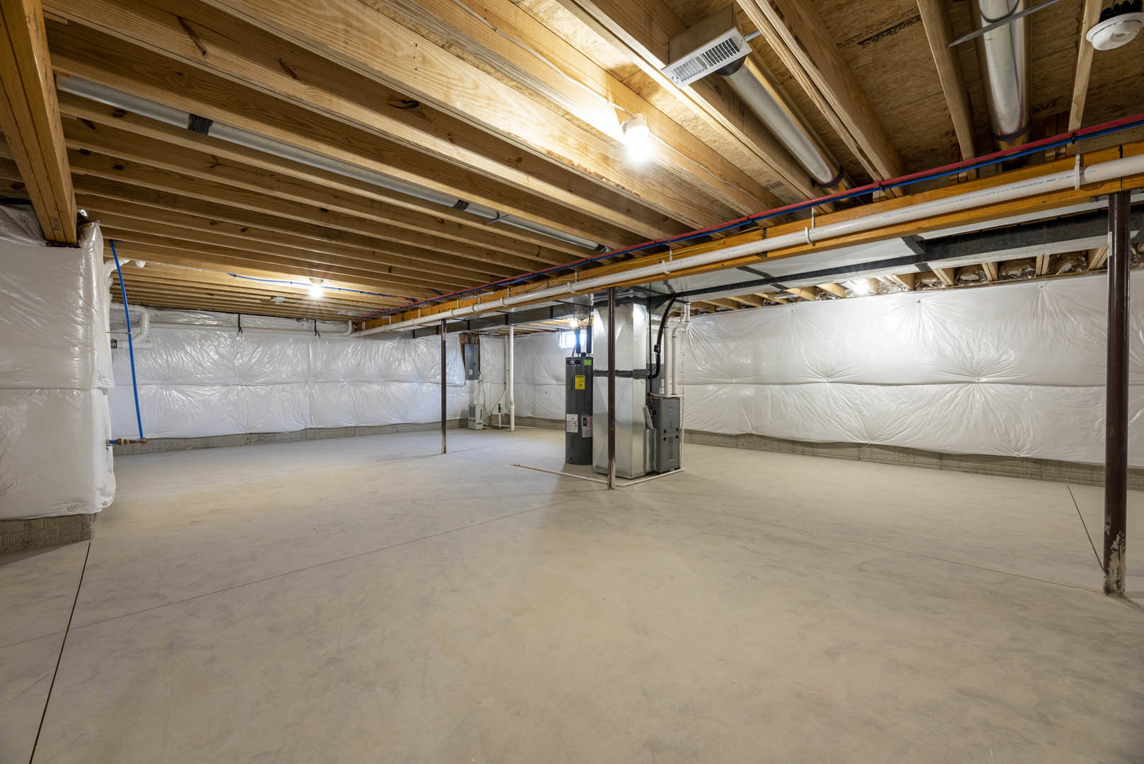 Spacious basement with exposed wood ceiling beams, white walls, steel support pole, metal utility box, and visible pipes along the ceiling.
