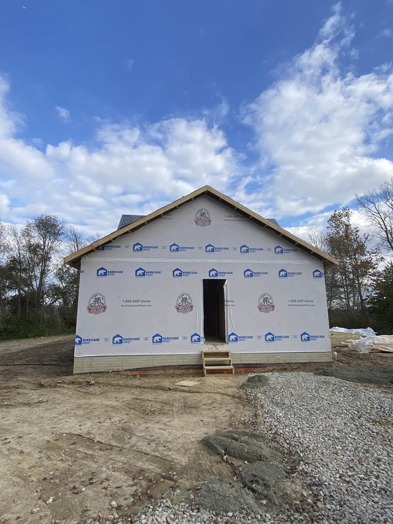 Partially built house with exposed wooden framing, open doorway, gravel foundation, and unfinished roof under blue sky with scattered clouds