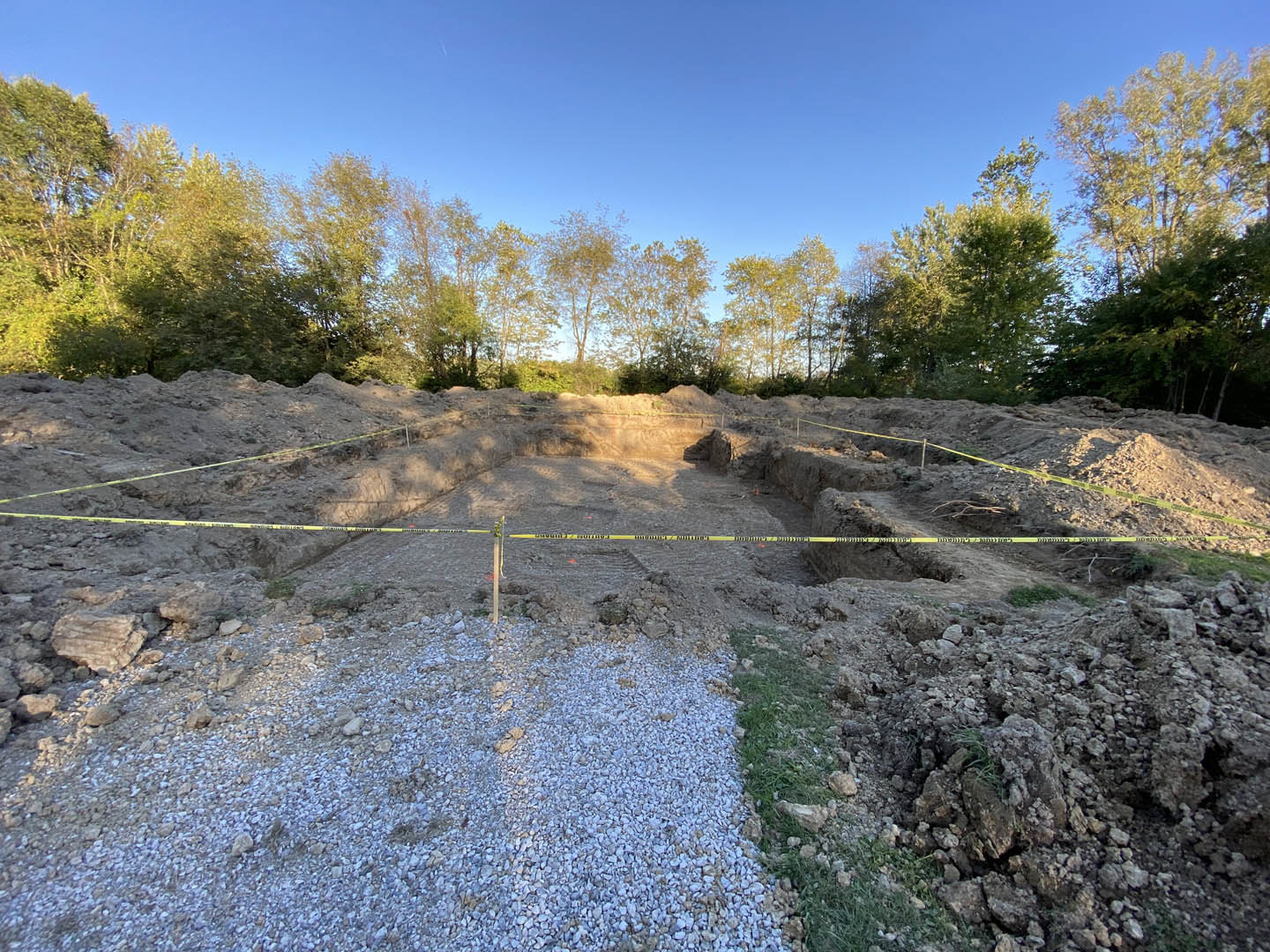 Rocky construction site bordered by yellow caution tape, with scattered stones and a sign in foreground, dense group of trees and blue sky in background