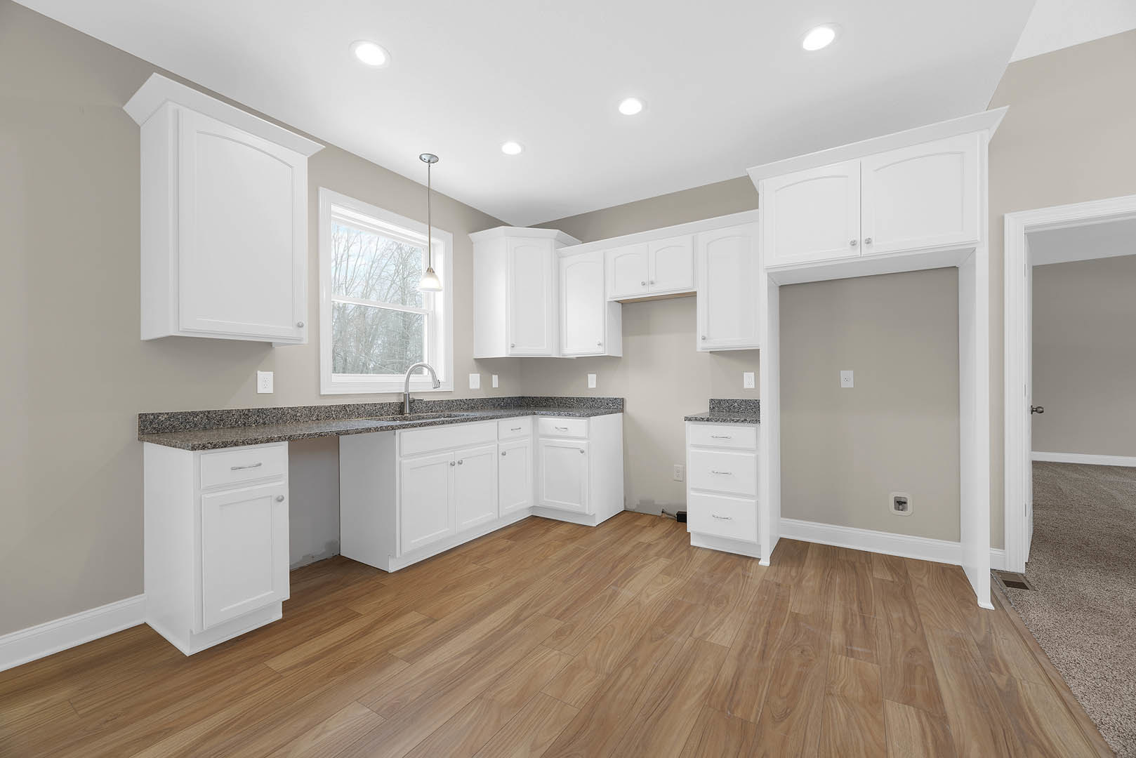 White kitchen cabinets with silver handles, wood flooring, stainless steel faucet over a sink, white door in the corner, window letting in natural light.