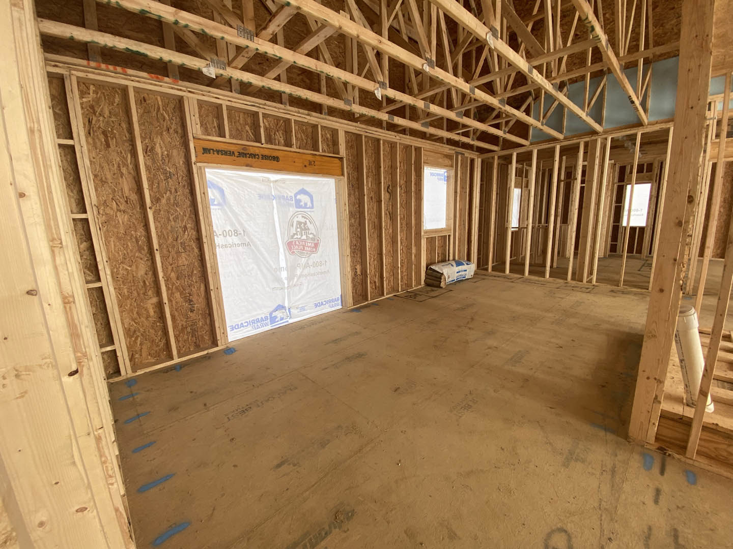 Unfinished room with exposed wooden framing, white plastic vapor barrier covering walls, wood beams on ceiling, window letting in natural light, bag of lumber on floor.