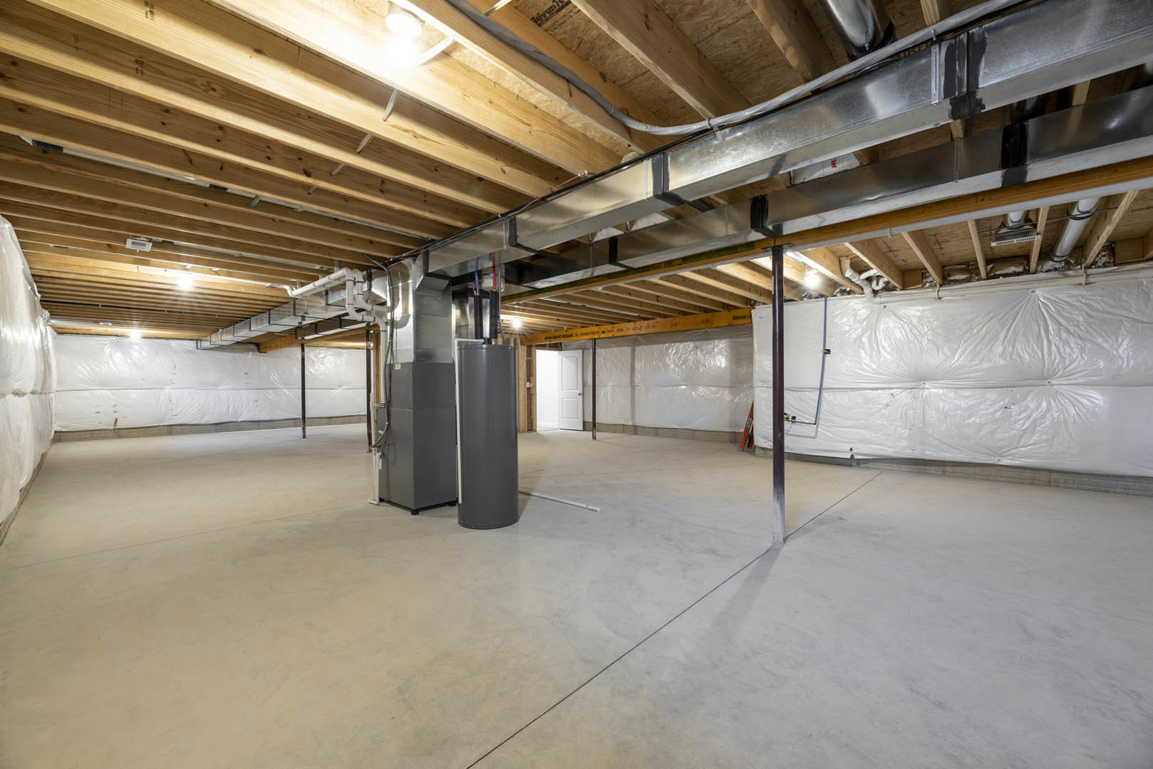 Basement room with exposed metal pole and ceiling pipes, white plastic wall covering, refrigerator, white floor with black line, and white door framed in wood.