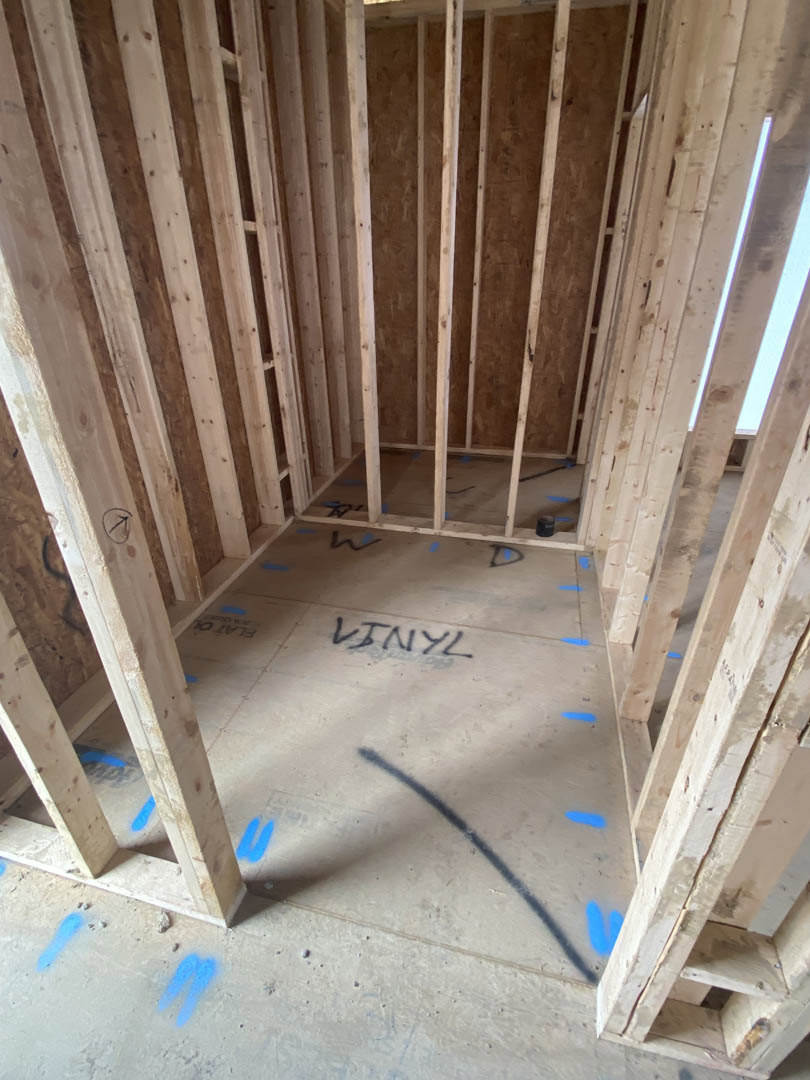Unfinished room with exposed wood framing, blue painter’s tape marking areas on subfloor, and construction materials scattered throughout
