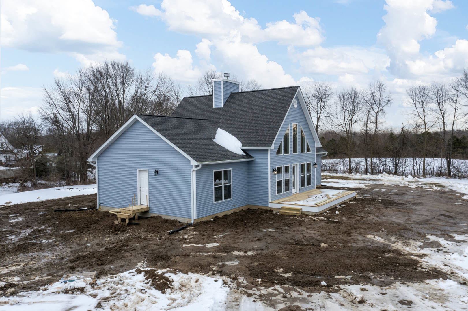 Two-story blue house under construction with snow on roof and patio, white-framed windows, chimney, muddy ground, bare trees in winter.
