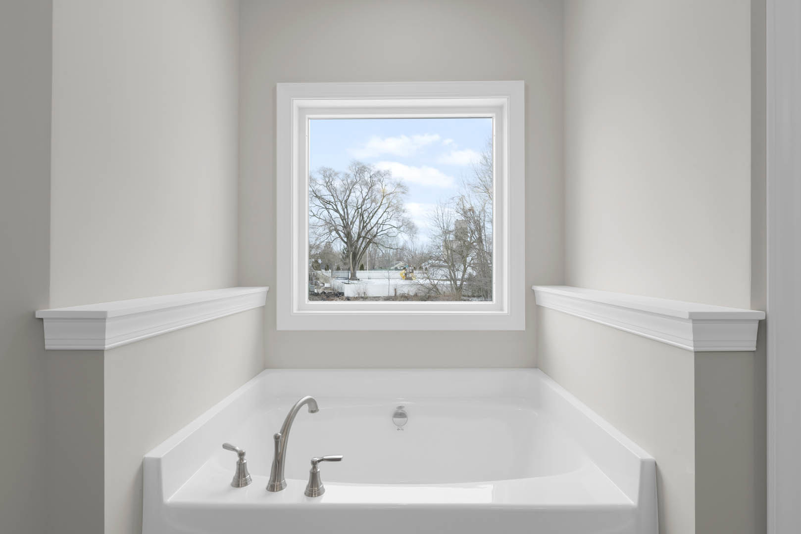 Freestanding white bathtub beneath large window with tree view, chrome faucet, white wall molding, and neutral bathroom finishes