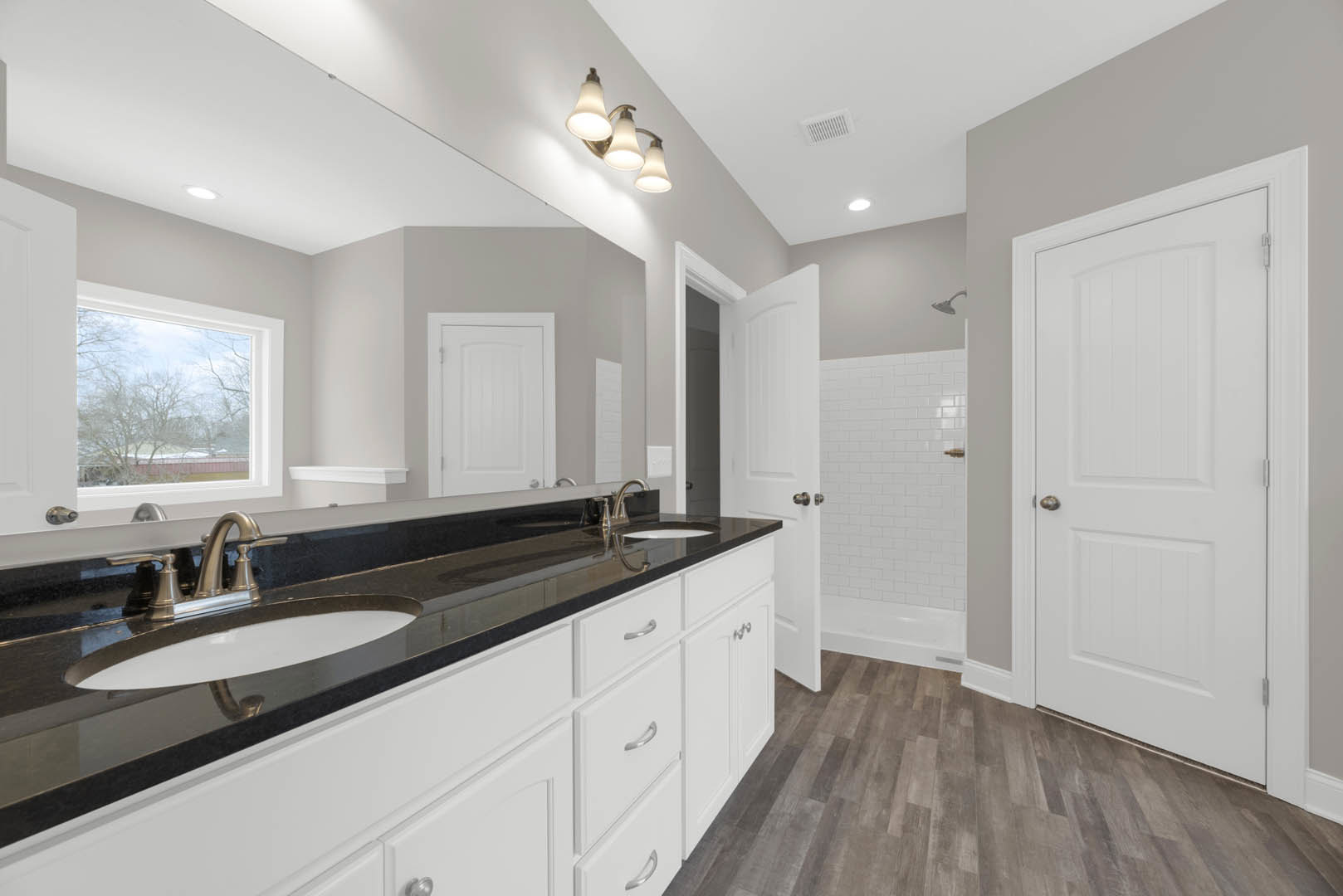Bathroom with two white sinks set in a stone countertop, wood plank flooring, white cabinetry, silver faucets, and a window overlooking a house and trees.