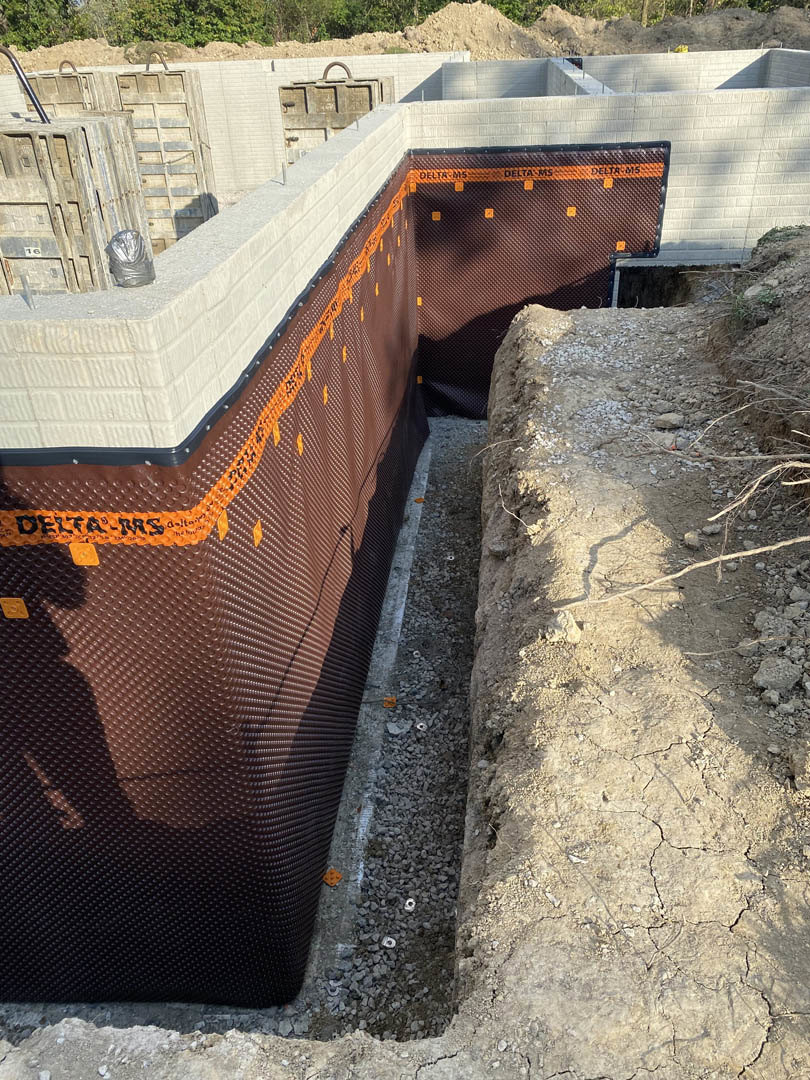 Concrete foundation slab bordered by brown soil, orange construction tape, and scattered piles of dirt, with metal crates stacked nearby and trees in the background.
