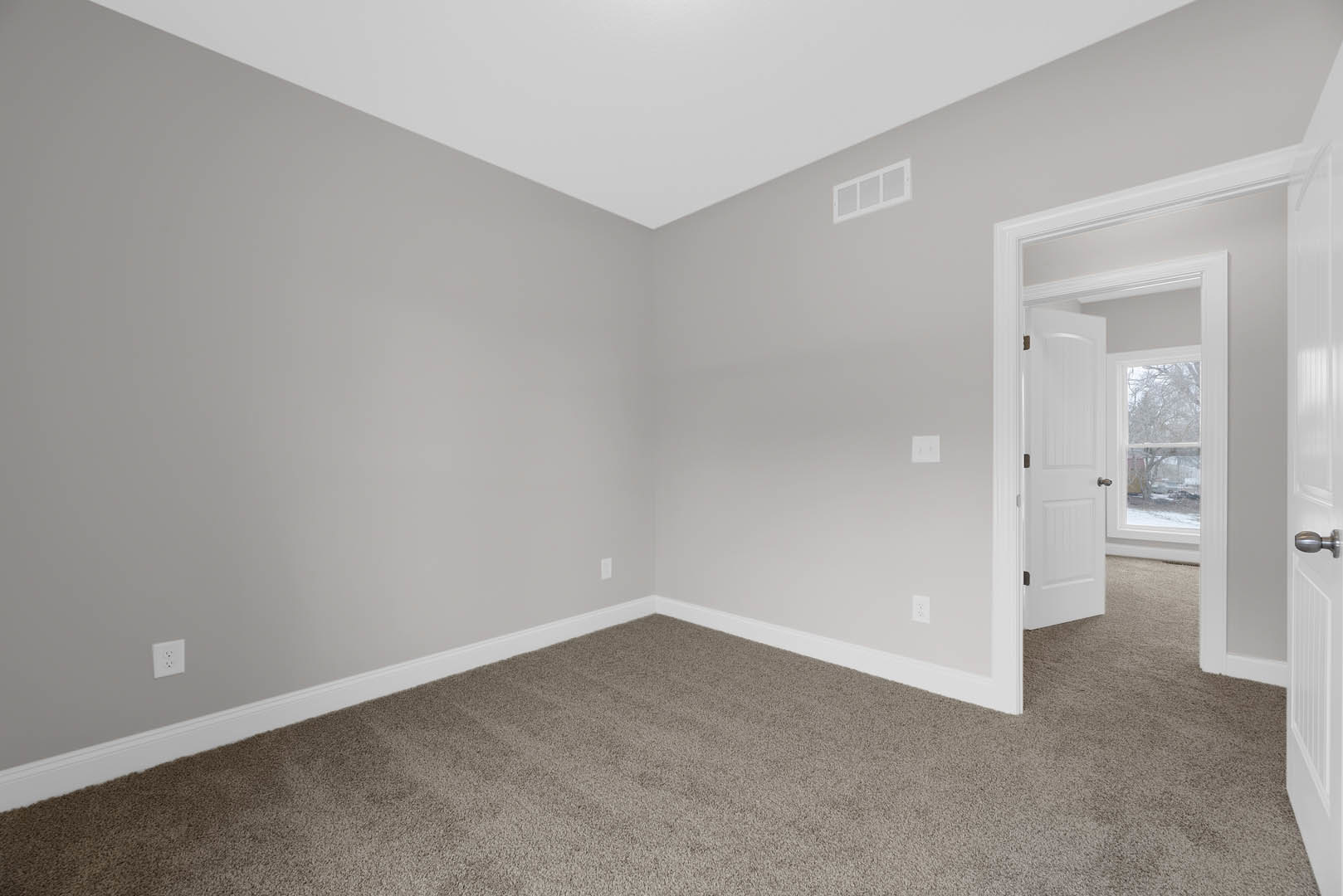 White door with silver doorknob open to a room featuring white walls, square white tiles, white electrical outlet, and light-colored carpet flooring under a cloudy sky.