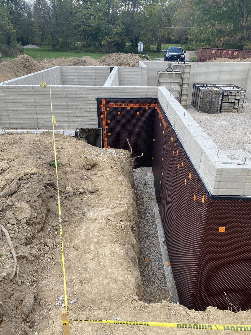 Black pickup truck parked beside stacked pallets at residential construction site, unfinished foundation and wall visible, trees in background