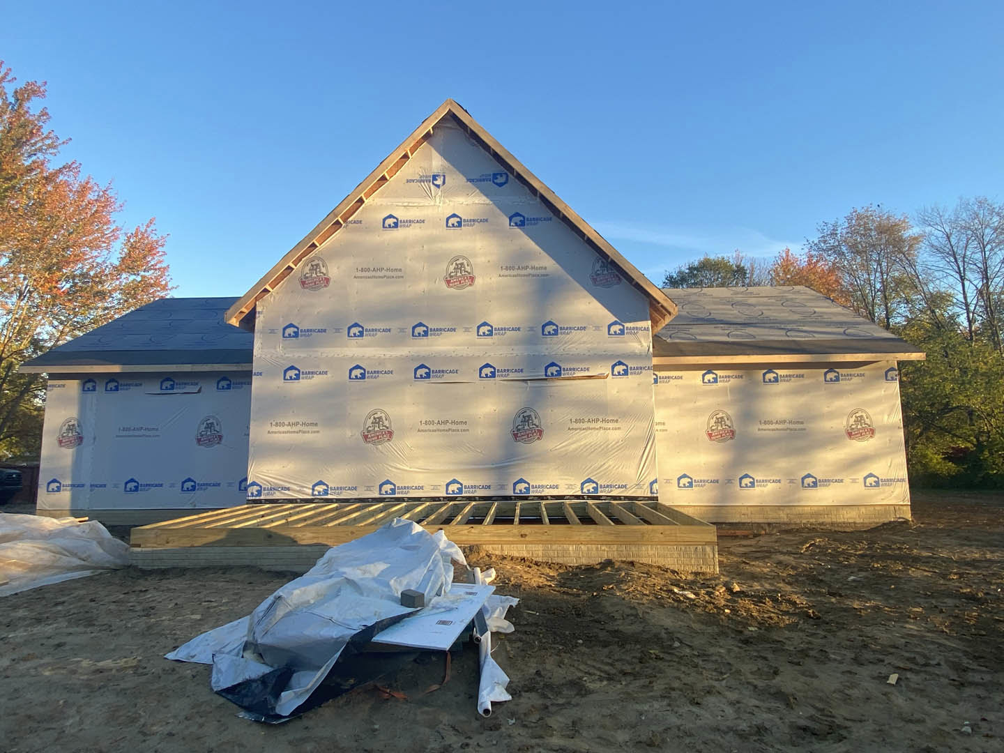 Partially built house with exposed framing, white plastic tarp covering materials on the ground, construction logo visible on wall, surrounding trees and blue sky in background
