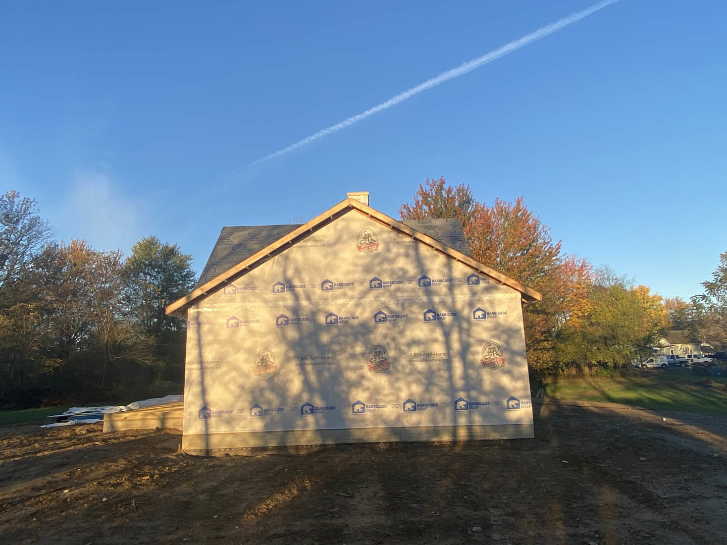 Partially built house framed with exposed wood, surrounded by trees under a clear blue sky, construction materials scattered on grassy lot