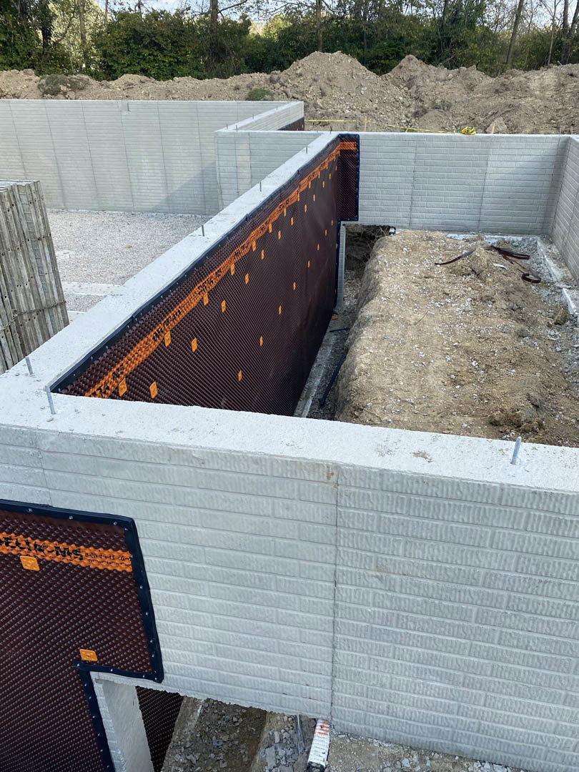 Concrete wall with orange and black tape, large pile of dirt, exposed hole in the ground, outdoor construction site with trees and plants in background