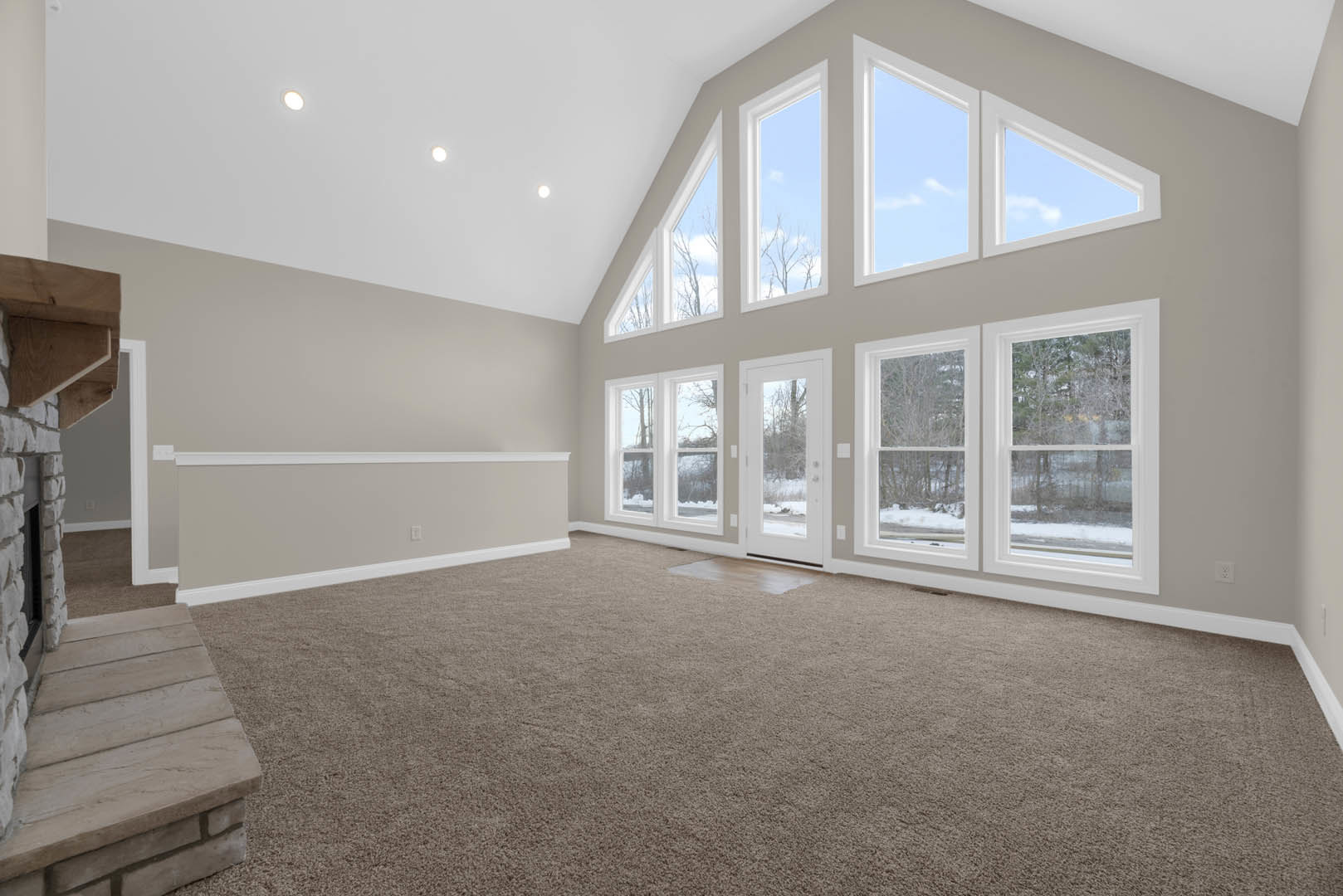 Carpeted living space featuring a large window with blue sky views, stone steps leading to a door, and molding accents; door opens to snowy forest scenery.