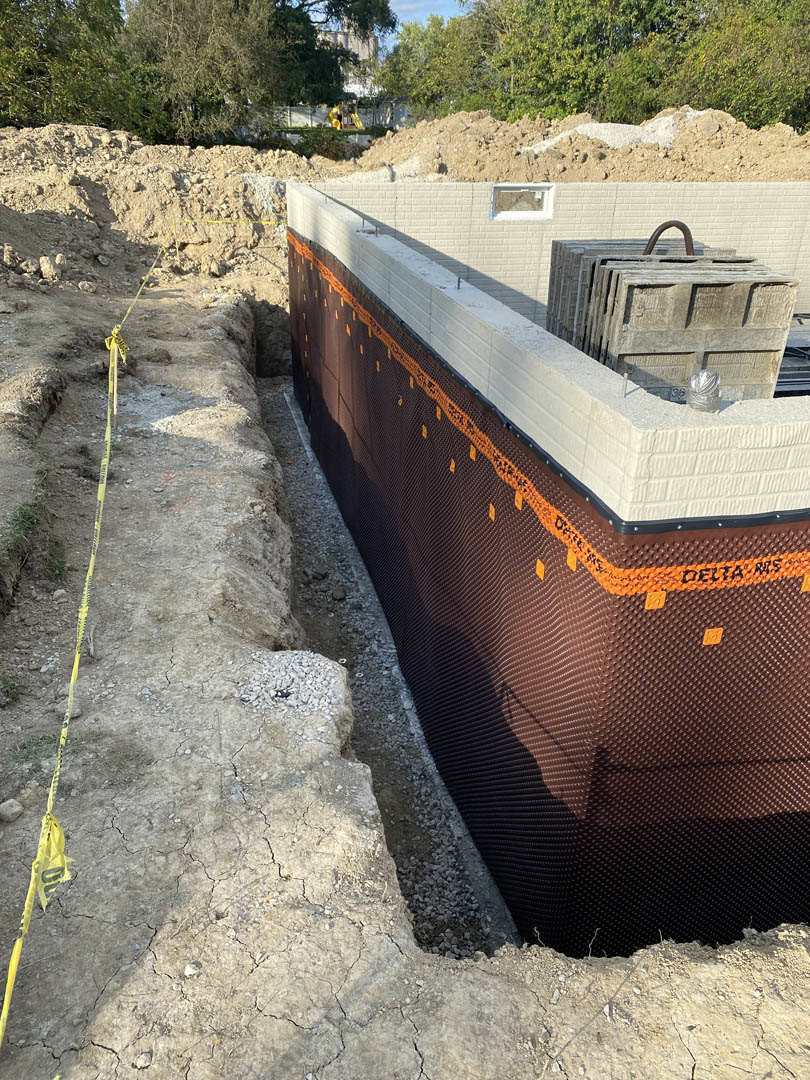 Concrete foundation with stacked blocks, dirt ground marked by yellow tape, construction materials and containers scattered on site, trees and sky in background