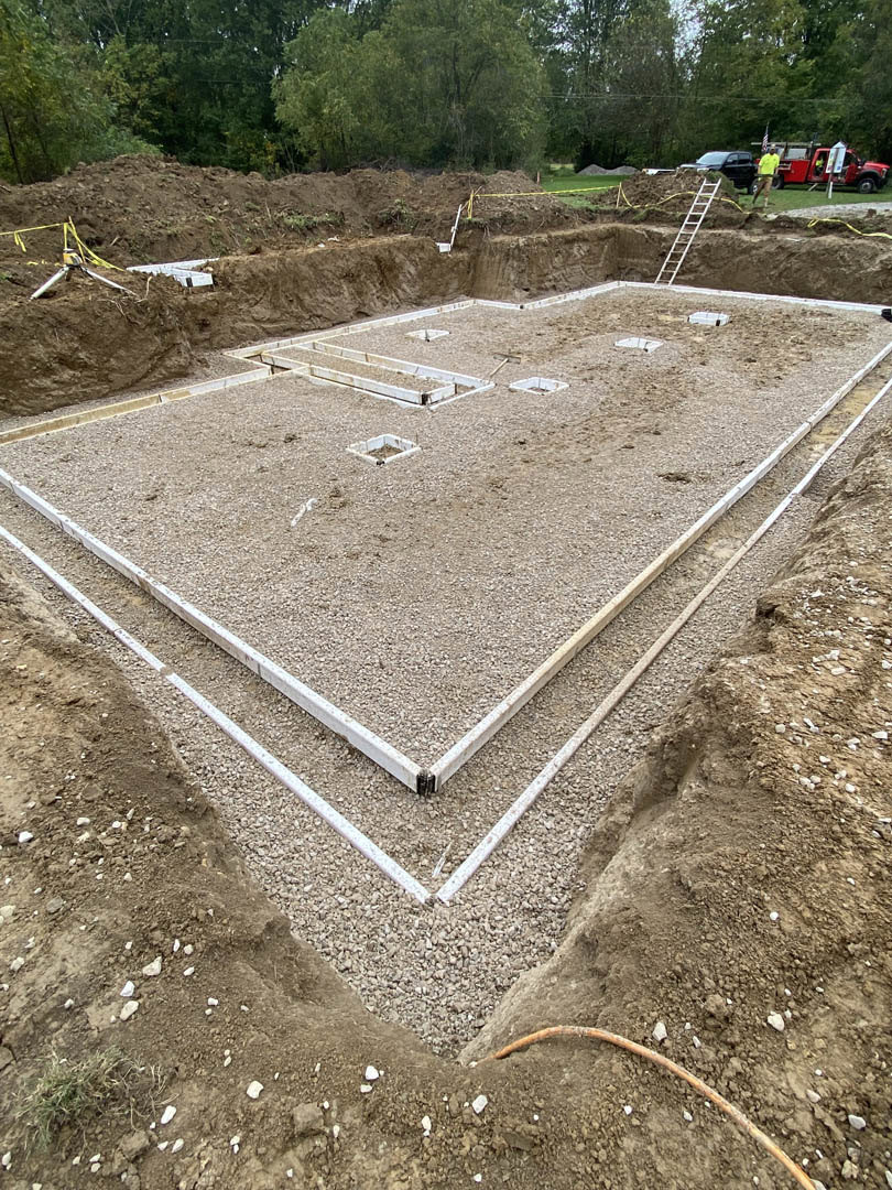 Framed foundation on a dirt construction site with workers, red truck, exposed pipe, ladder on slope, and surrounding trees