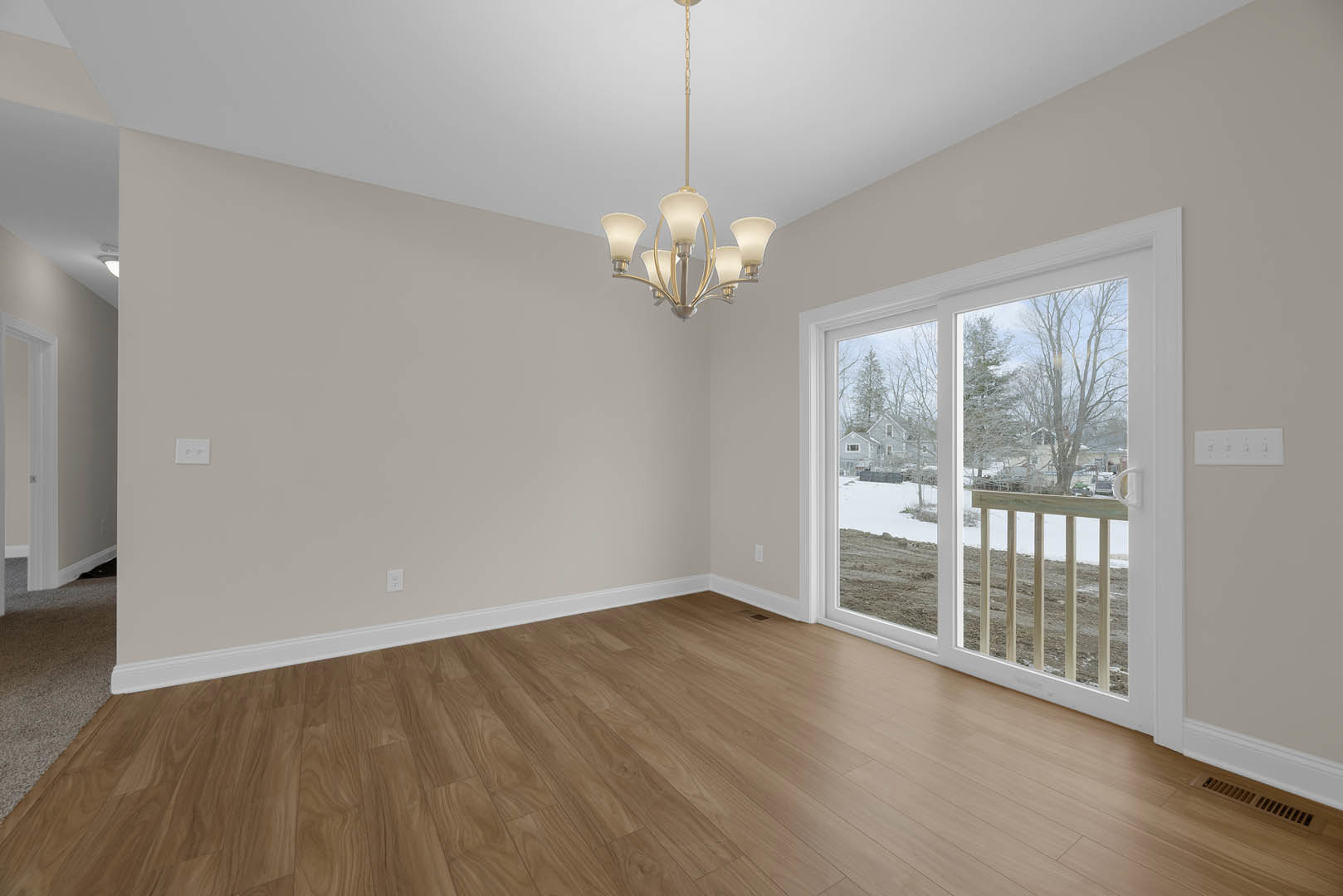 Open living space with hardwood floors, modern chandelier hanging from the ceiling, white stair railing, and large sliding glass door overlooking a snowy backyard.