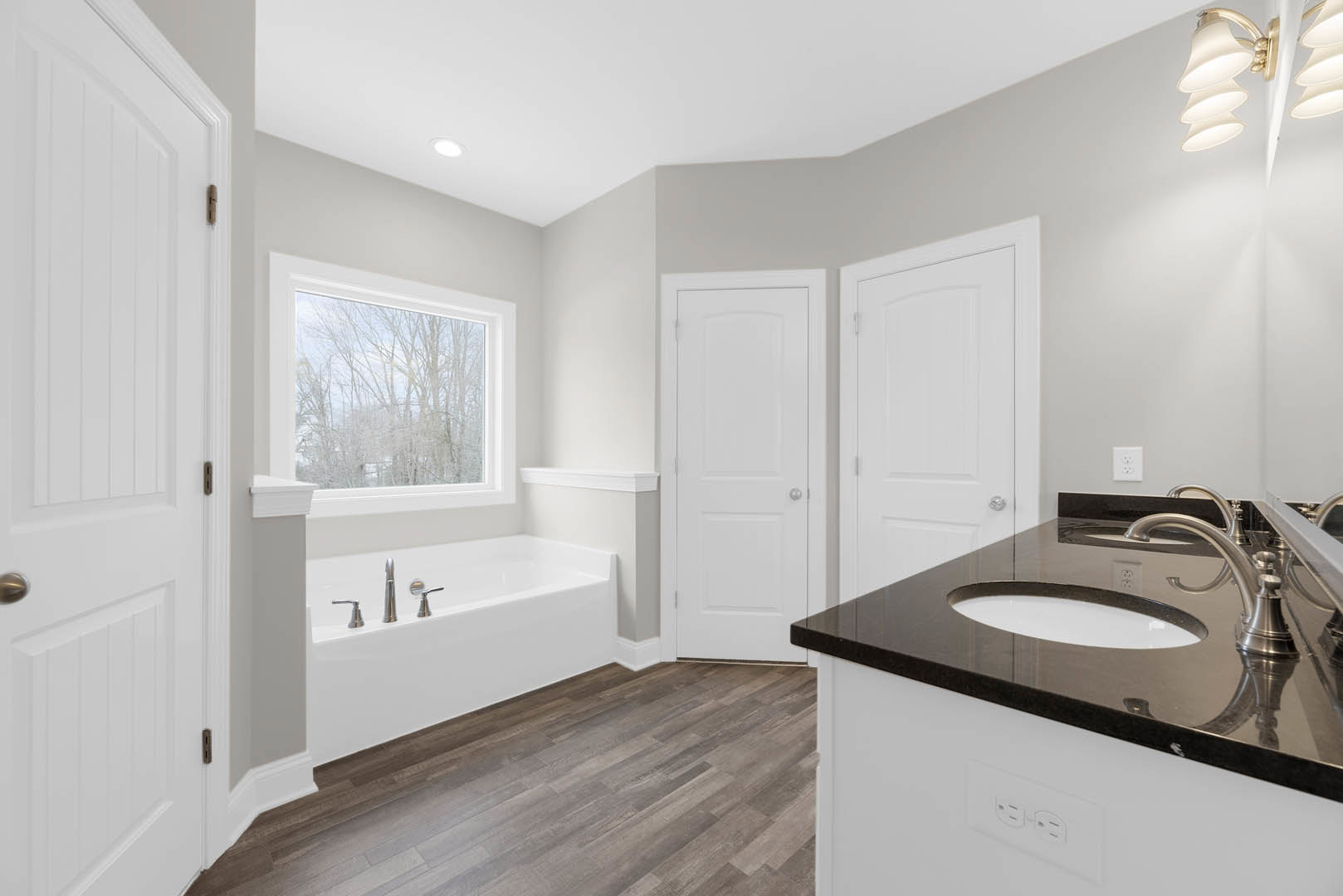 Bathroom with freestanding white tub, rectangular sink on stone countertop, tiled walls, window overlooking trees, white door with silver knob, modern lamp fixture