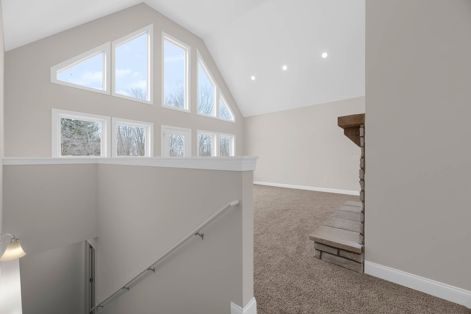 Curved staircase with carpeted steps and stone ledge, wall of tall windows letting in natural light, white pipe with metal hooks, tree and blue sky visible through windows