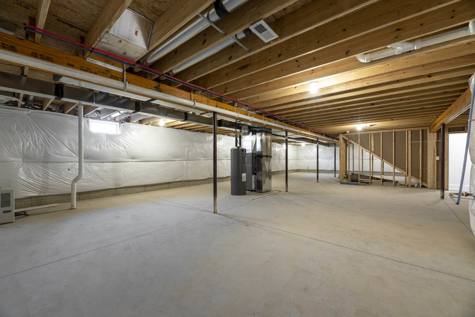 Basement with white painted walls, exposed pipes, concrete floor, wood ceiling beams, black steel support pillar with white markings, vent, metal utility box, and white plastic