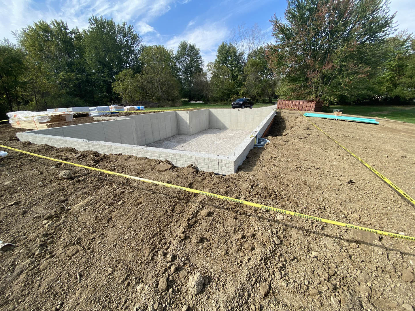 Concrete foundation surrounded by piles of dirt and rocks, parked car in front, trees and cloudy sky in background