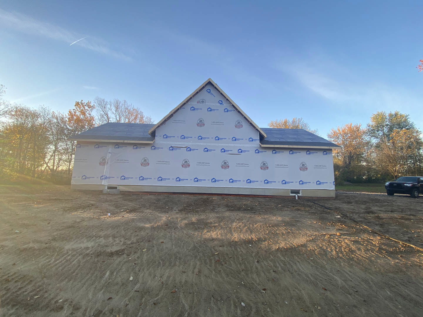 Modern house under construction with white plastic covering the roof, black car parked on paved road, sandy lot with construction sign, surrounded by trees under clear blue sky