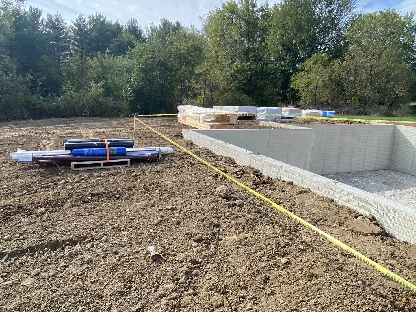 Concrete foundation with exposed pipes and yellow tape measure, surrounded by dirt and construction materials; trees and sky visible in background