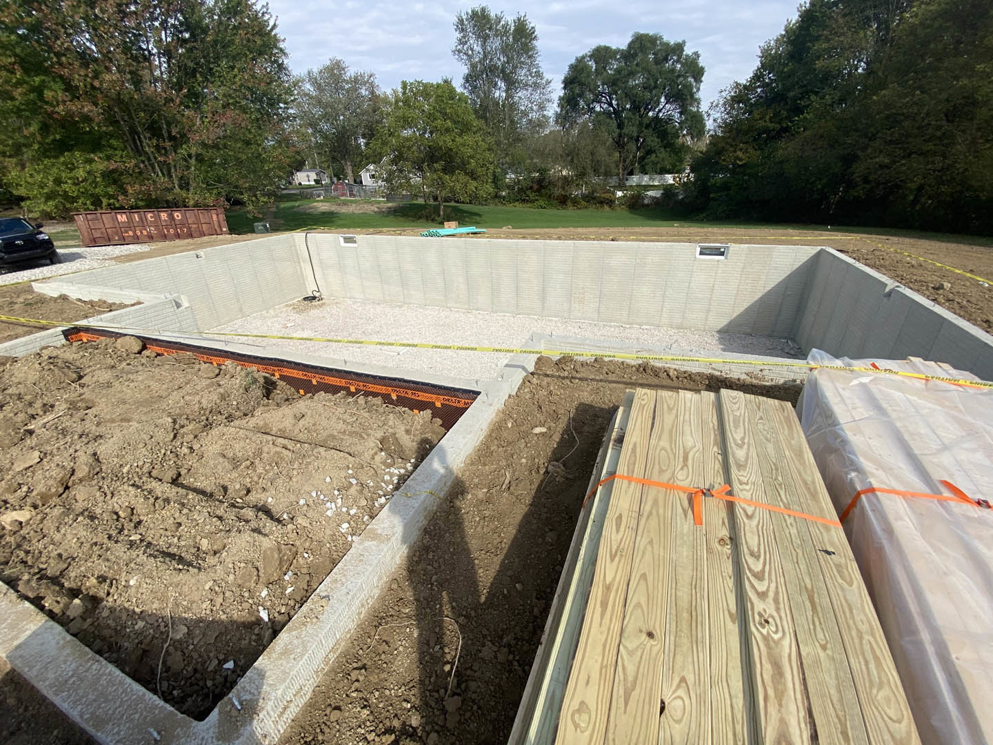 Residential construction site with exposed foundation, stacks of lumber marked with orange ribbon, piles of dirt, yellow caution tape, and scattered wooden planks on bare ground