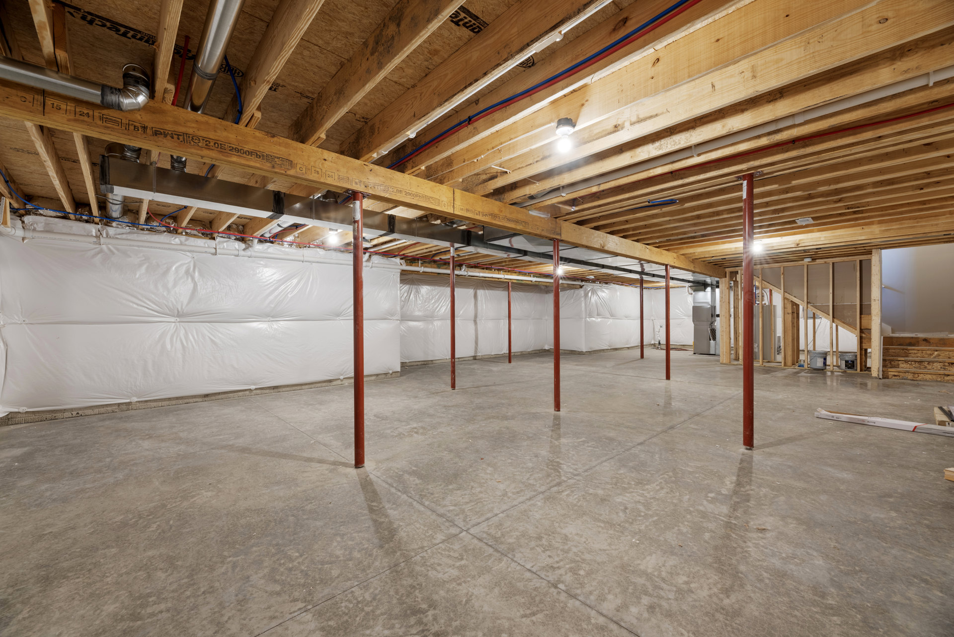 Exposed wooden ceiling beams, white walls with plastic covering, grey concrete floor with central line, red support poles, single light bulb hanging from ceiling